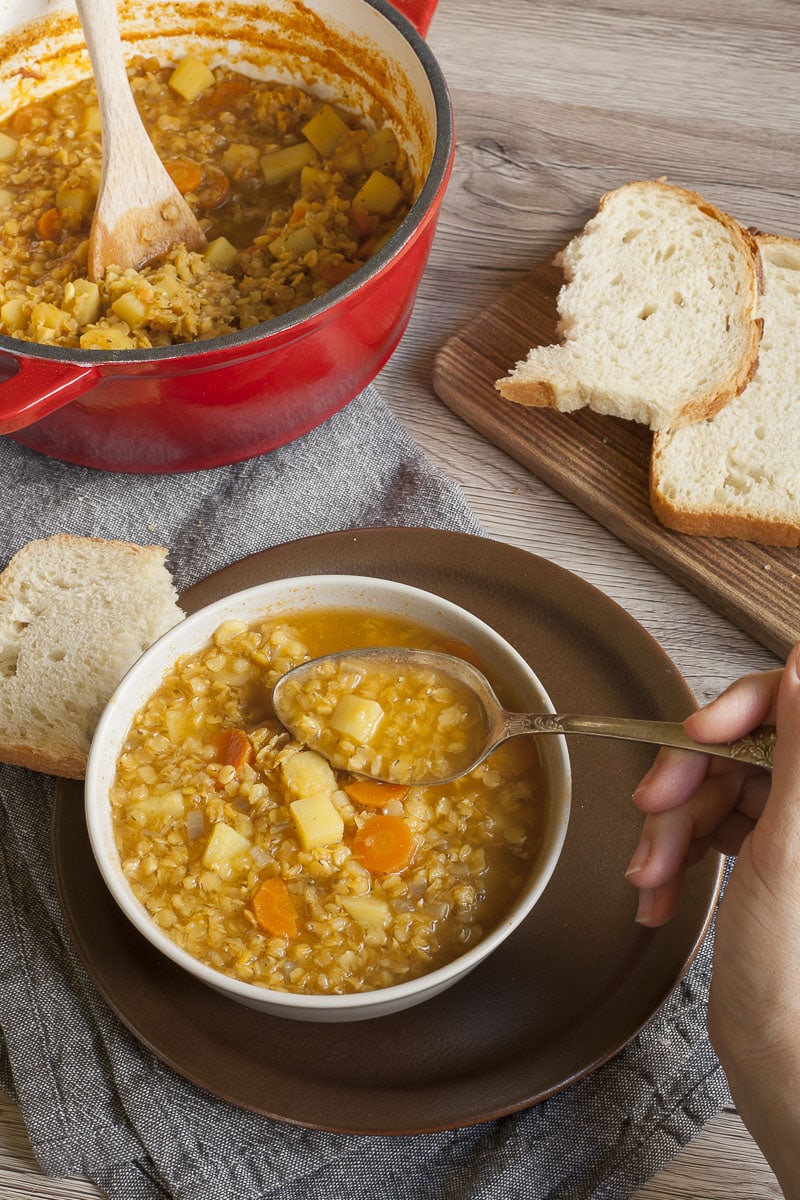 White bowl with red lentil soup and a hand is taking a spoonful. The remaining soup is in the red Dutch Oven in the background