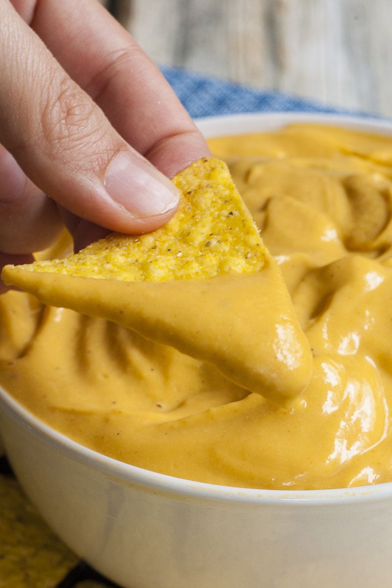 White bowl of dairy-free nut-free cheese sauce and a hand dips a nacho chips in it