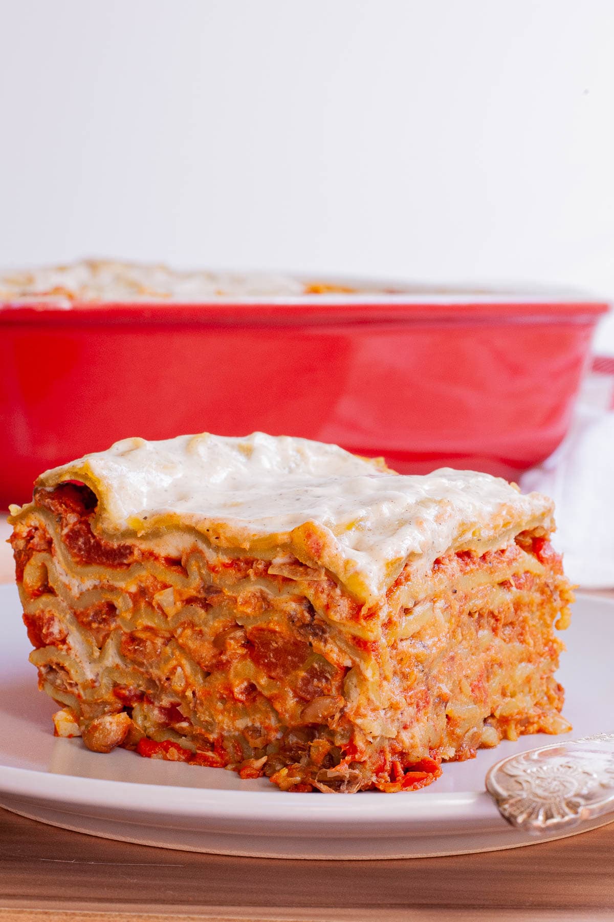A slice of layered lasagna is served on a plate. A red casserole dish with the remaining lasagna is right behind it.