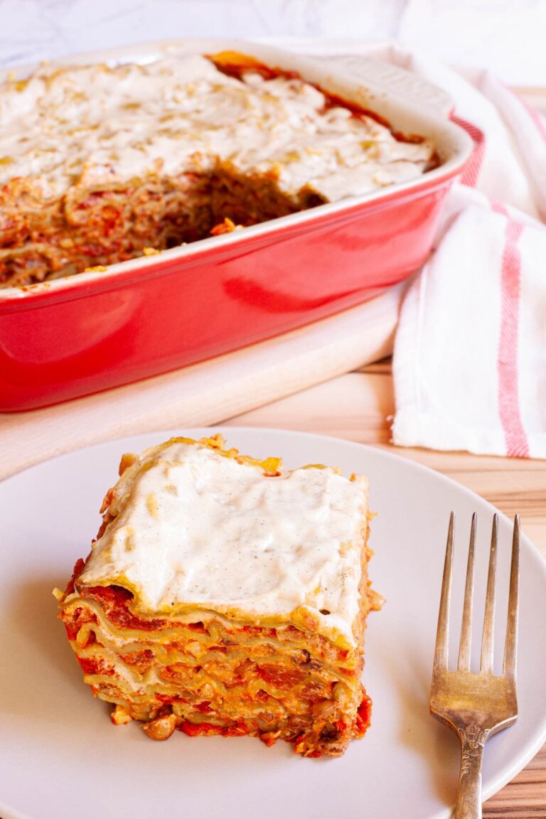 A slice of layered lasagna is served on a plate. A red casserole dish with the remaining lasagna is right behind it.