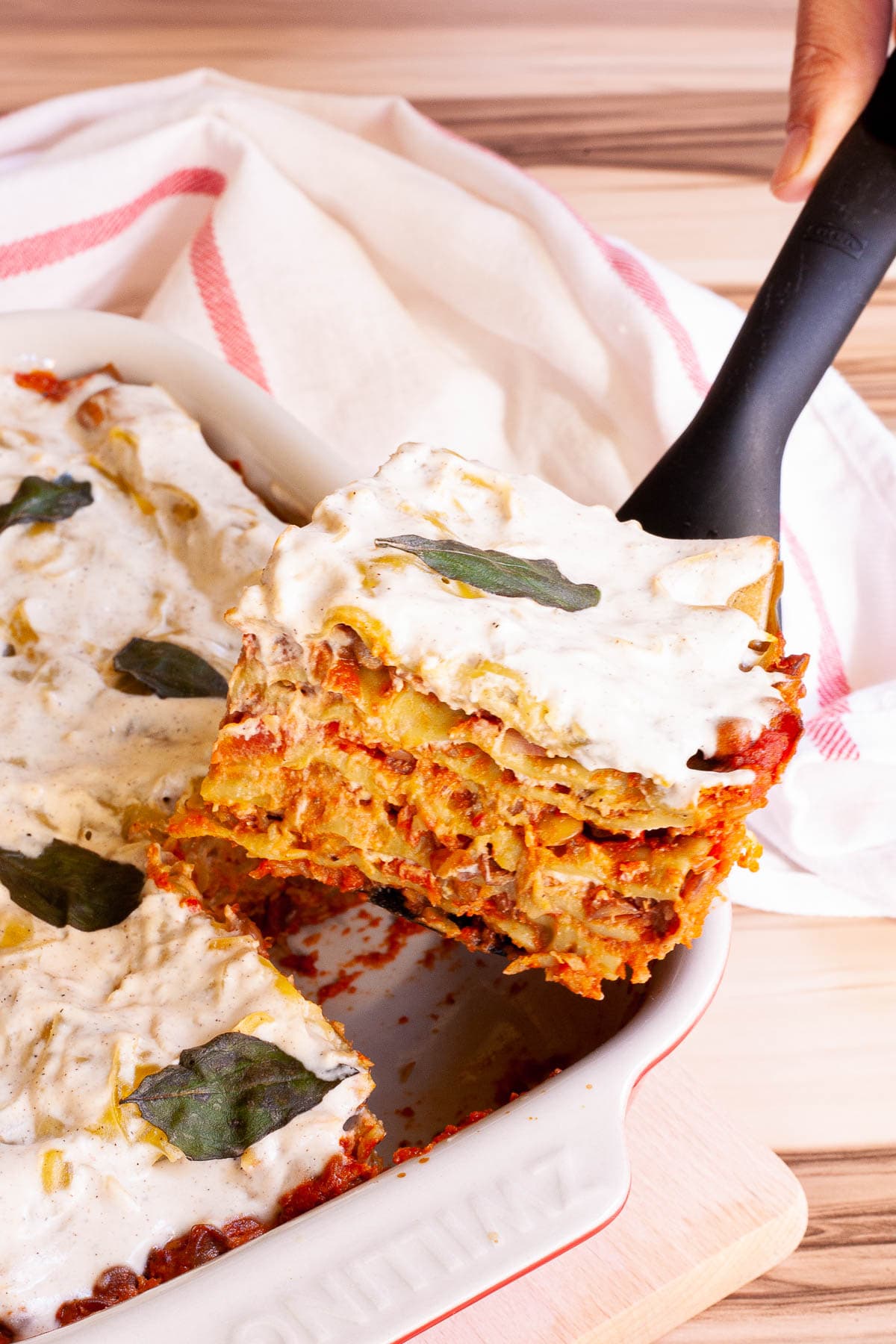 A hand is holding a black slotted turner and lifting a large slice of layered lasagna out of the casserole.