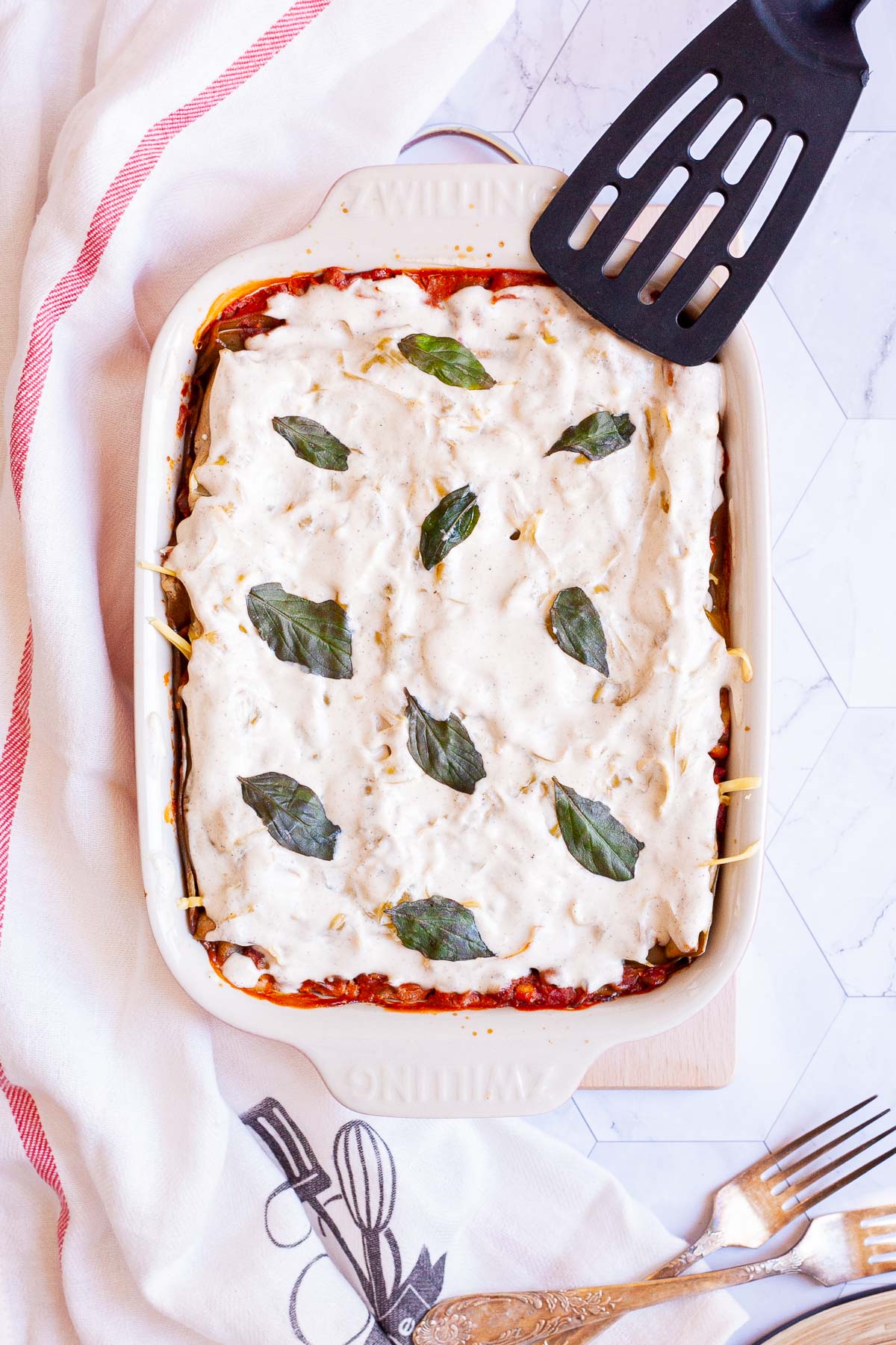 Rectangular casserole with a meal but only the white sauce on top is visible and some basil leaves. A slotted turner is placed next to it.