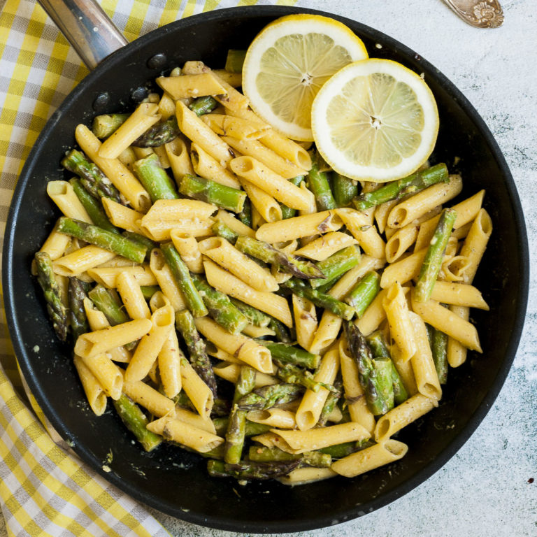 Frying pan from above with penne pasta, asparagus pieces and 2 lemon slices.