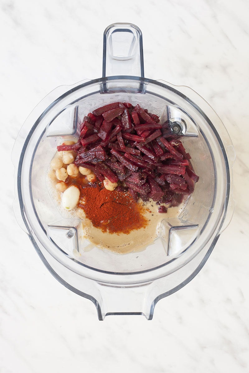 A blender with roasted beet pieces, chickpeas, tahini, lemon juice, garlic clove, and paprika powder.