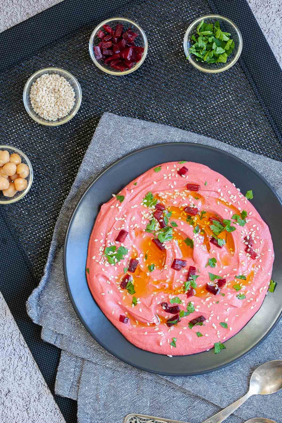 Pink hummus on a black plate, sprinkled with freshly chopped parsley, sesame seeds and small roasted beet pieces. Small glass bowls with chickpeas, sesame seeds, roasted diced beet and chopped parsley are around it.