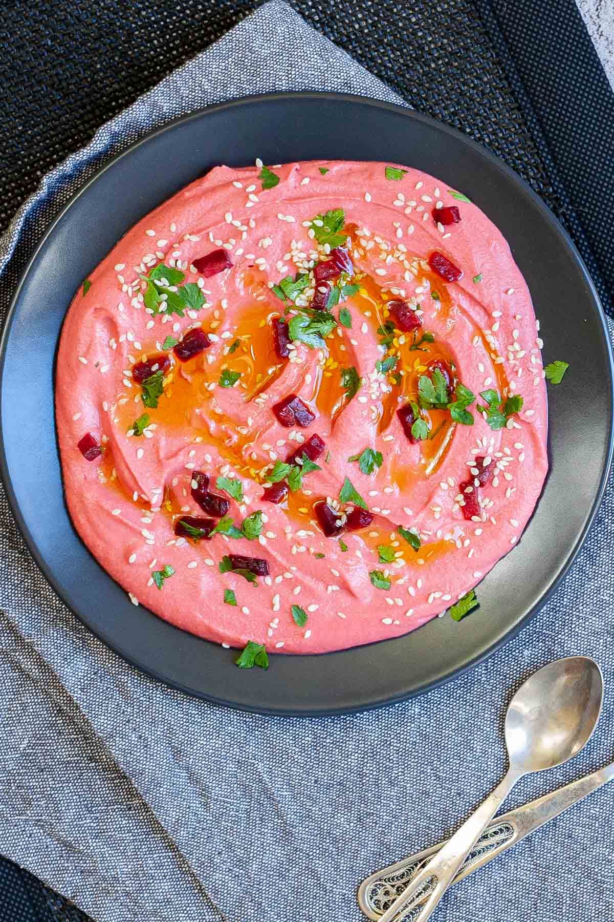 Pink hummus on a black plate, sprinkled with freshly chopped parsley, sesame seeds and small roasted beet pieces.