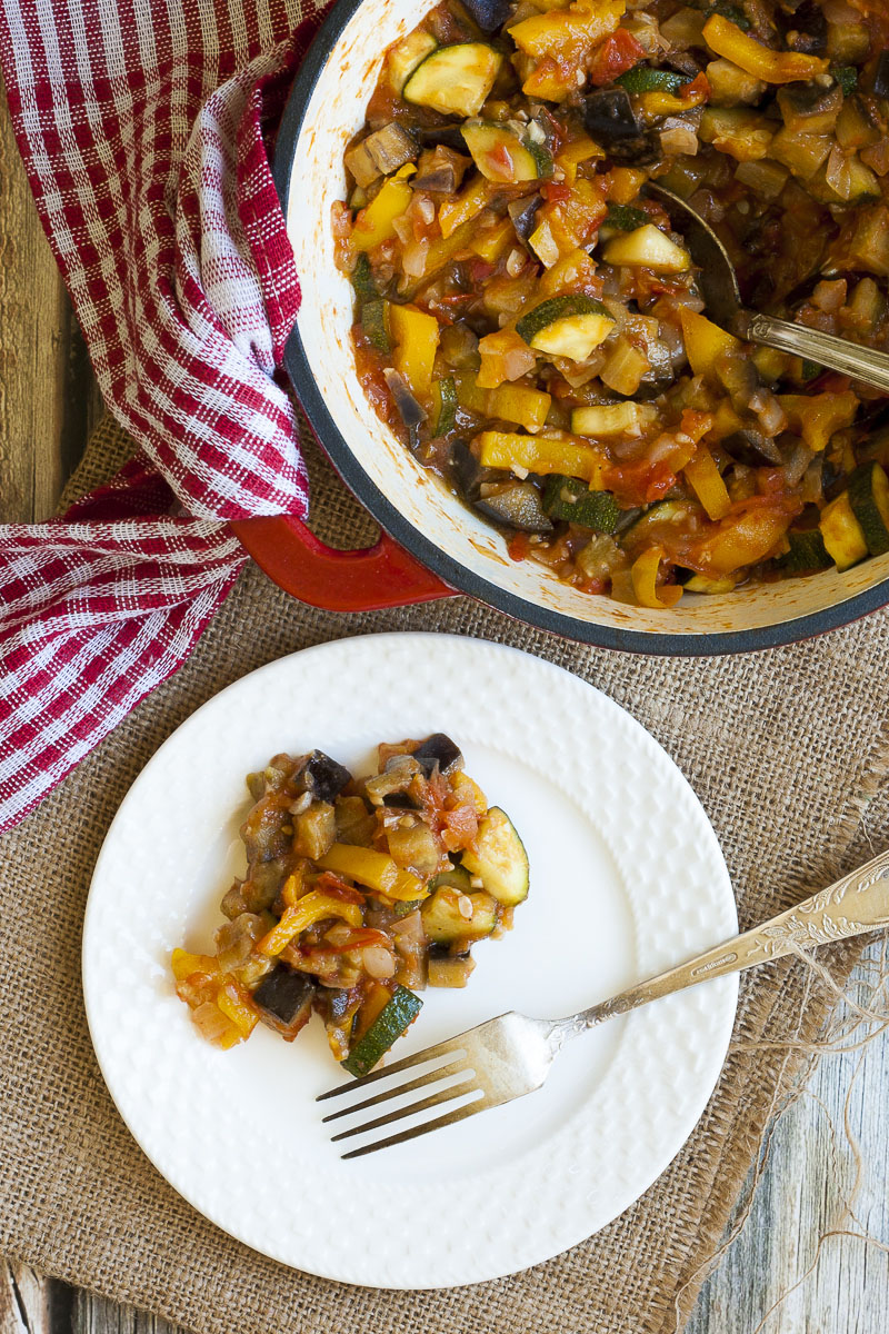 White plate from above with a small portion of served ratatouille - red, yellow, green, brown chunks. Next to it is a large white pot with the remaining veggie chunks. 