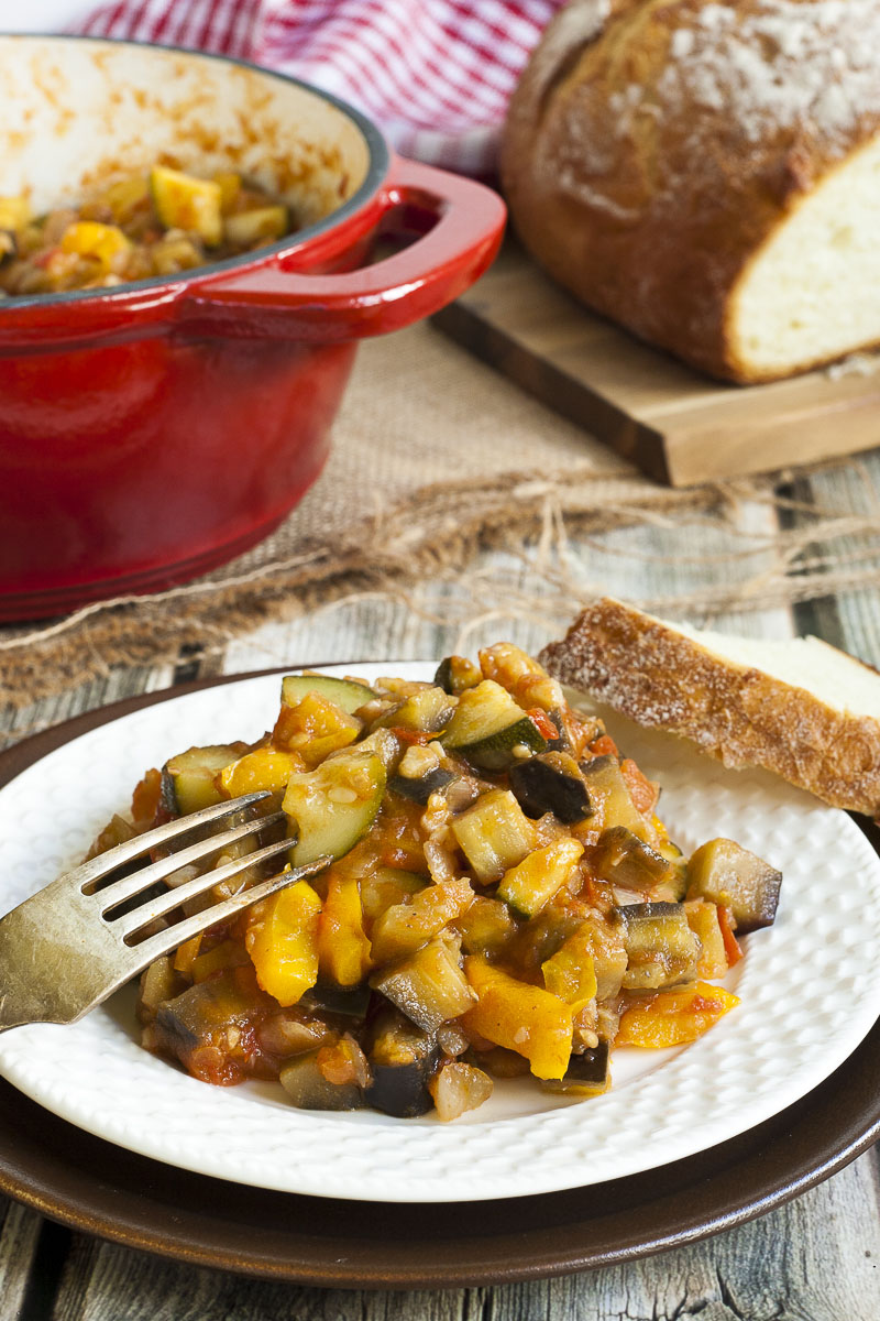 White plate up close with a small portion of ratatouille - red, yellow, green, brown chunks. Behind it there is is a large red pot with the remaining veggie chunks and a loaf of crusty bread on the side.