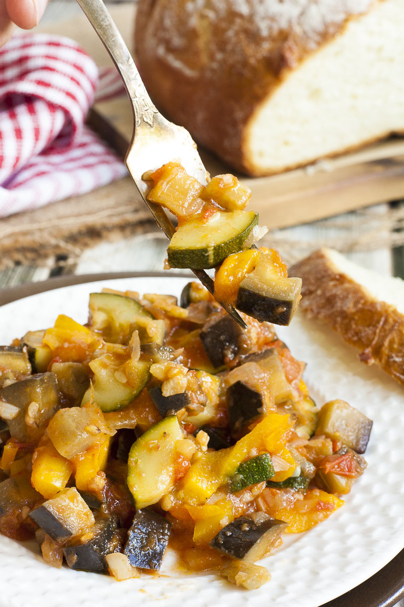 White plate with a small portion of ratatouille up close - red, yellow, green, brown chunks. A fork is taking a mouthful. 