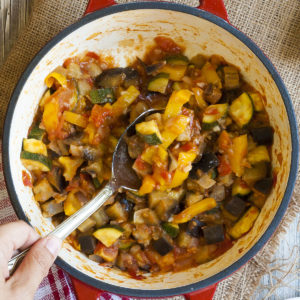 A red pot from above with veggie chunks of all colors: red, yellow, green, orange, brown. There is a serving spoon taking a portion from the middle.