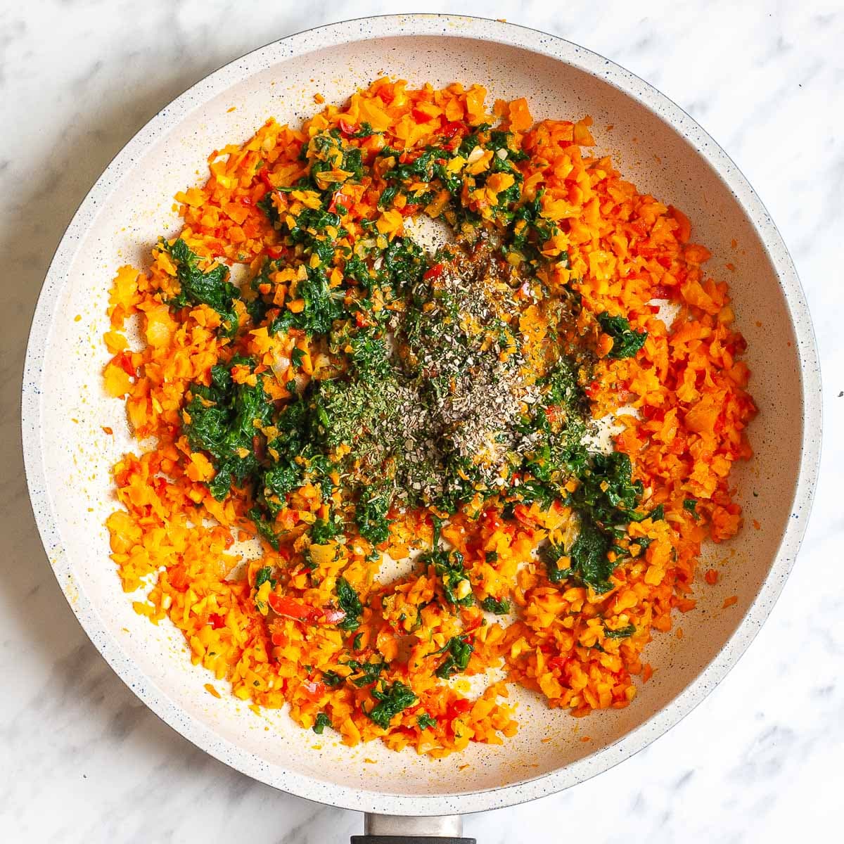 White frying pan with finely chopped carrots, onion, red pepper, and garlic pieces topped with spinach pieces and spices. 