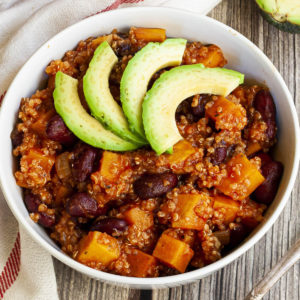 In a small white bowl orange cubes, dark brown beans, small white, yellow seeds with 3 slices of green avocado on top.