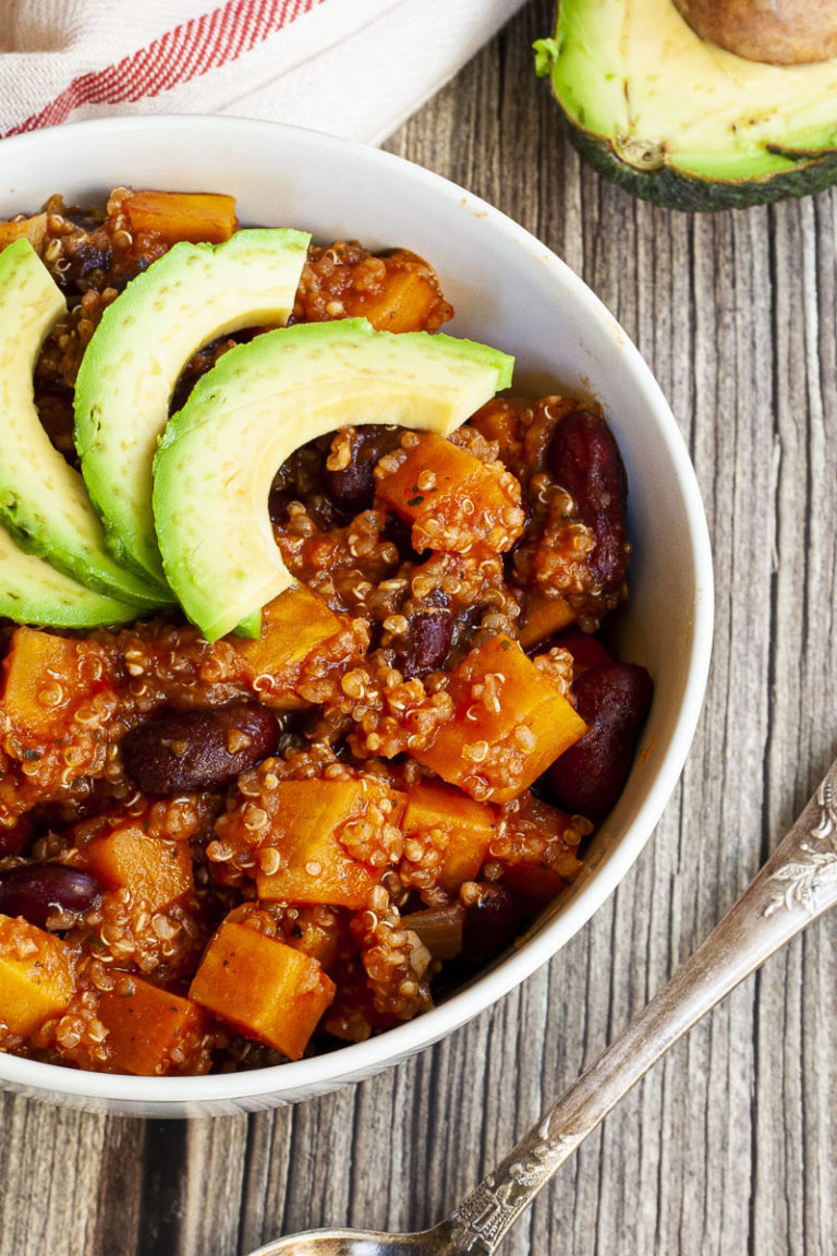 In a small white bowl orange cubes, dark brown beans, small white, yellow seeds with 3 slices of green avocado on top.