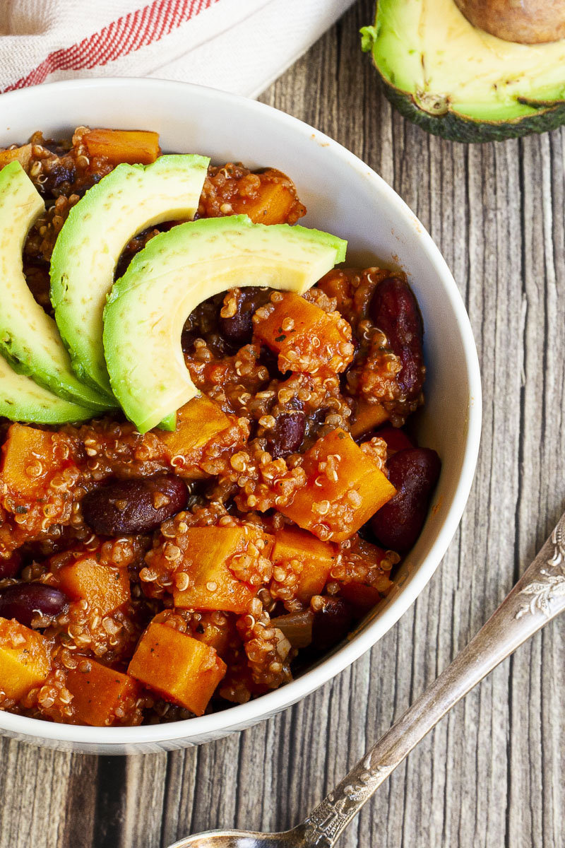 In a small white bowl orange cubes, dark brown beans, small white, yellow seeds with 3 slices of green avocado on top. 