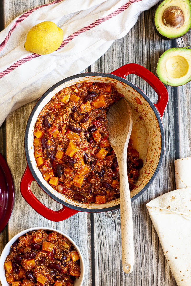 In a red dutch oven there are orange cubes, dark brown beans, small white, yellow seeds from above. There is a large wooden spoon as some of the chili has been already served. 