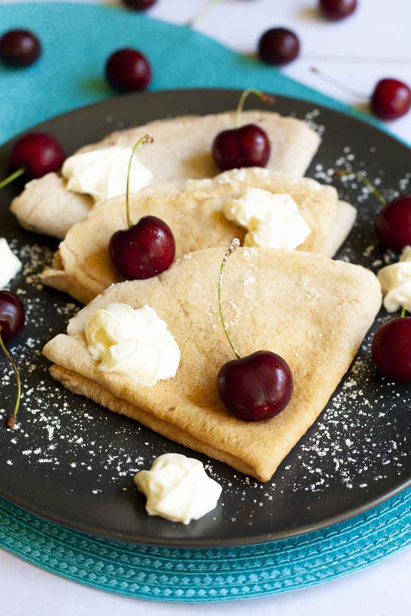 Black plate from top with 3 crepes folded as triangles with cherry and whipped cream scattered on top. 