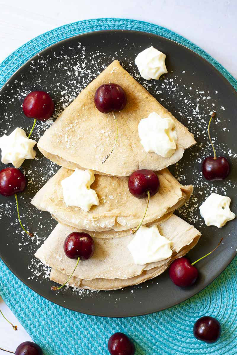 Black plate from top with 3 crepes folded as triangles with cherry and whipped cream scattered on top. 