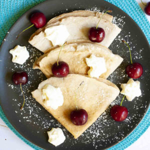 Black plate from top with 3 crepes folded as triangles with cherry and whipped cream scattered on top.