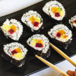 6 sushi rolls on a black plate. The rolls have black cover and white inside with purple yellow and green in the middle.