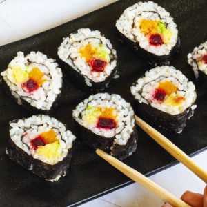 6 sushi rolls on a black plate. The rolls have black cover and white inside with purple yellow and green in the middle.