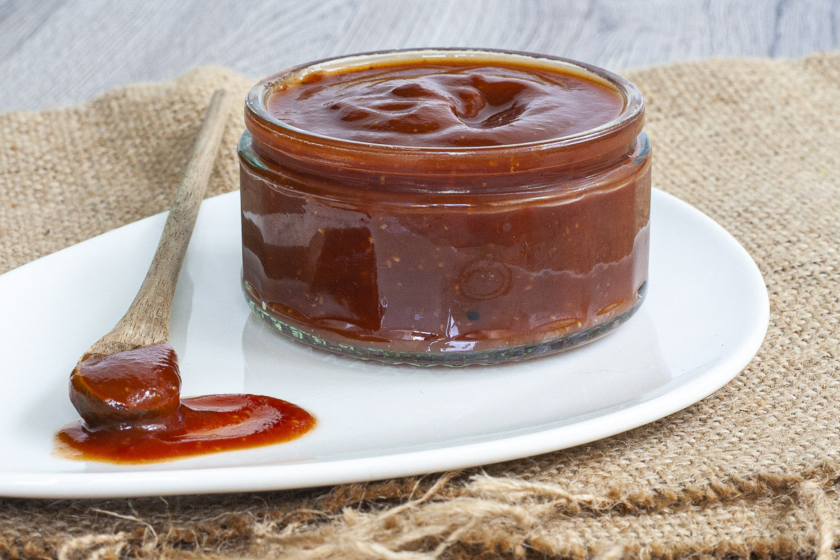 Small glass container full of red thick sauce. A small wooden spoon is laid next to it on a white plate dripping with red sauce. 