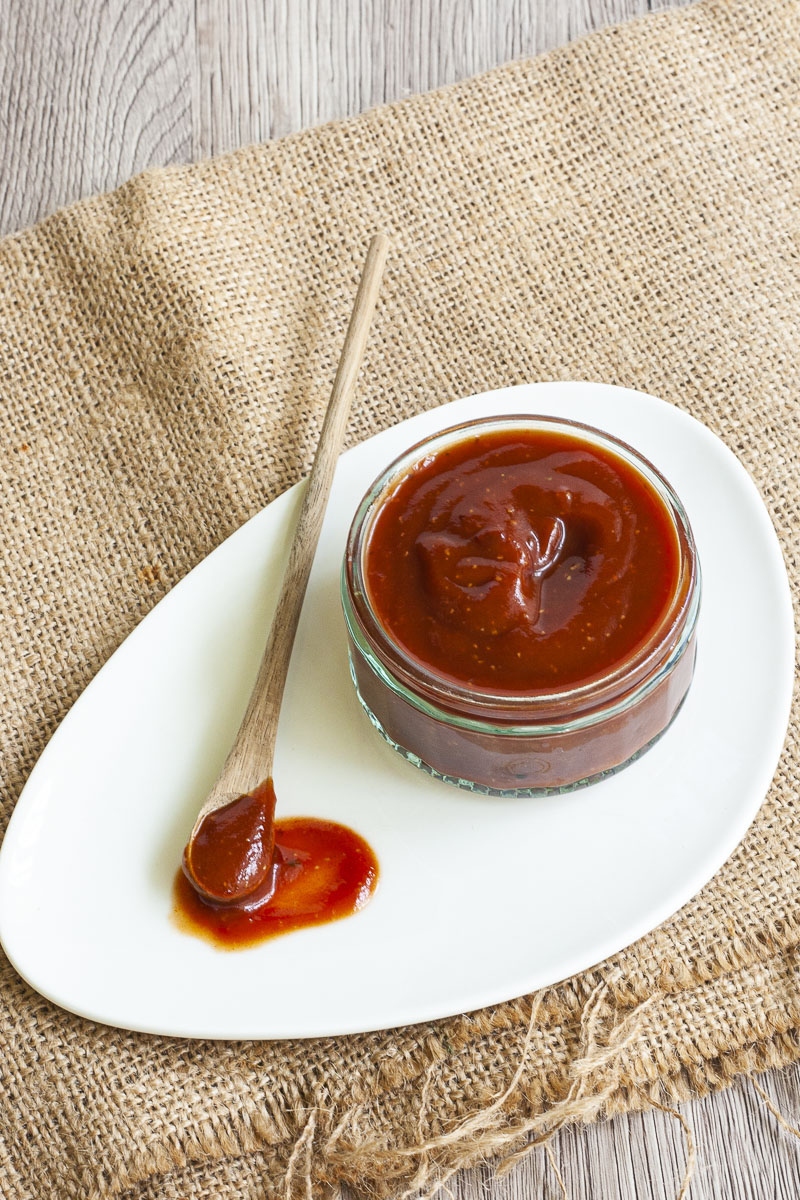 Small glass container from above full of red thick sauce. A small wooden spoon is laid next to it on a white plate dripping with red sauce. 