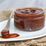 Small glass container up close full of red thick sauce. A small wooden spoon is laid next to it on a white plate dripping with red sauce.