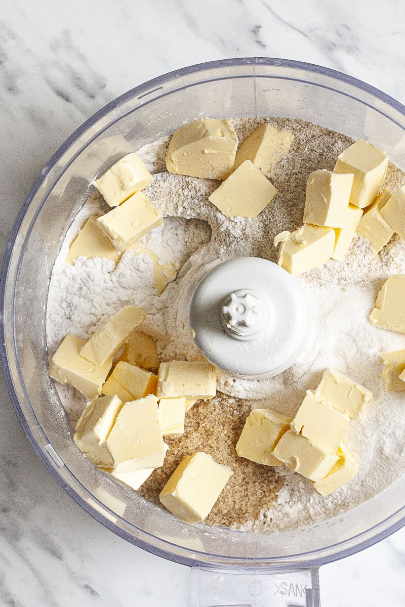 Open food processor from above with the ingredients of vegan gluten-free pie crust, different white and brown powders and yellow cubes of butter