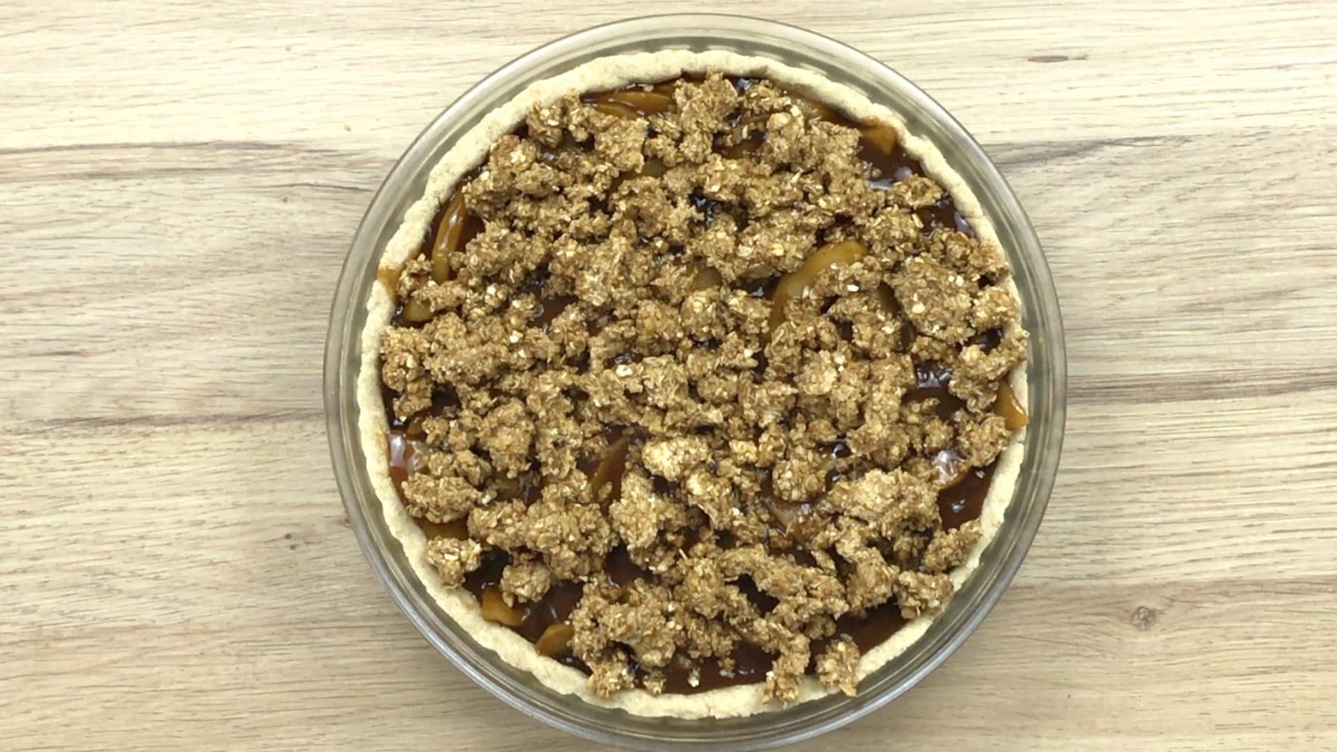 A pie crust in a glass pie pan filled with apple slices in caramel sauce topped with oat crisp.