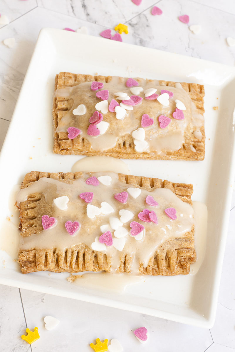 2 pop tarts, rectangle-shaped pillow-like cookies on a white plate. Both has white glaze and white and pink small hearts on the top.