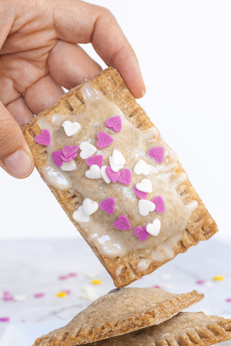 A hand is holding a pop tart, a rectangle-shaped pillow-like cookie with white glaze on top and pink and white small hearts