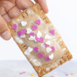 A hand is holding a pop tart, a rectangle-shaped pillow-like cookie with white glaze on top and pink and white small hearts