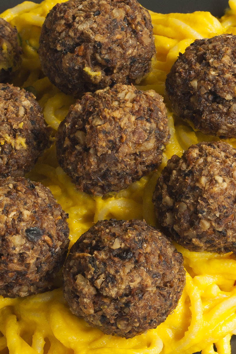 Several mushroom meatballs on top of spaghetti with yellow creamy sauce served on a black plate. A fork is taking on meatballs from the middle.