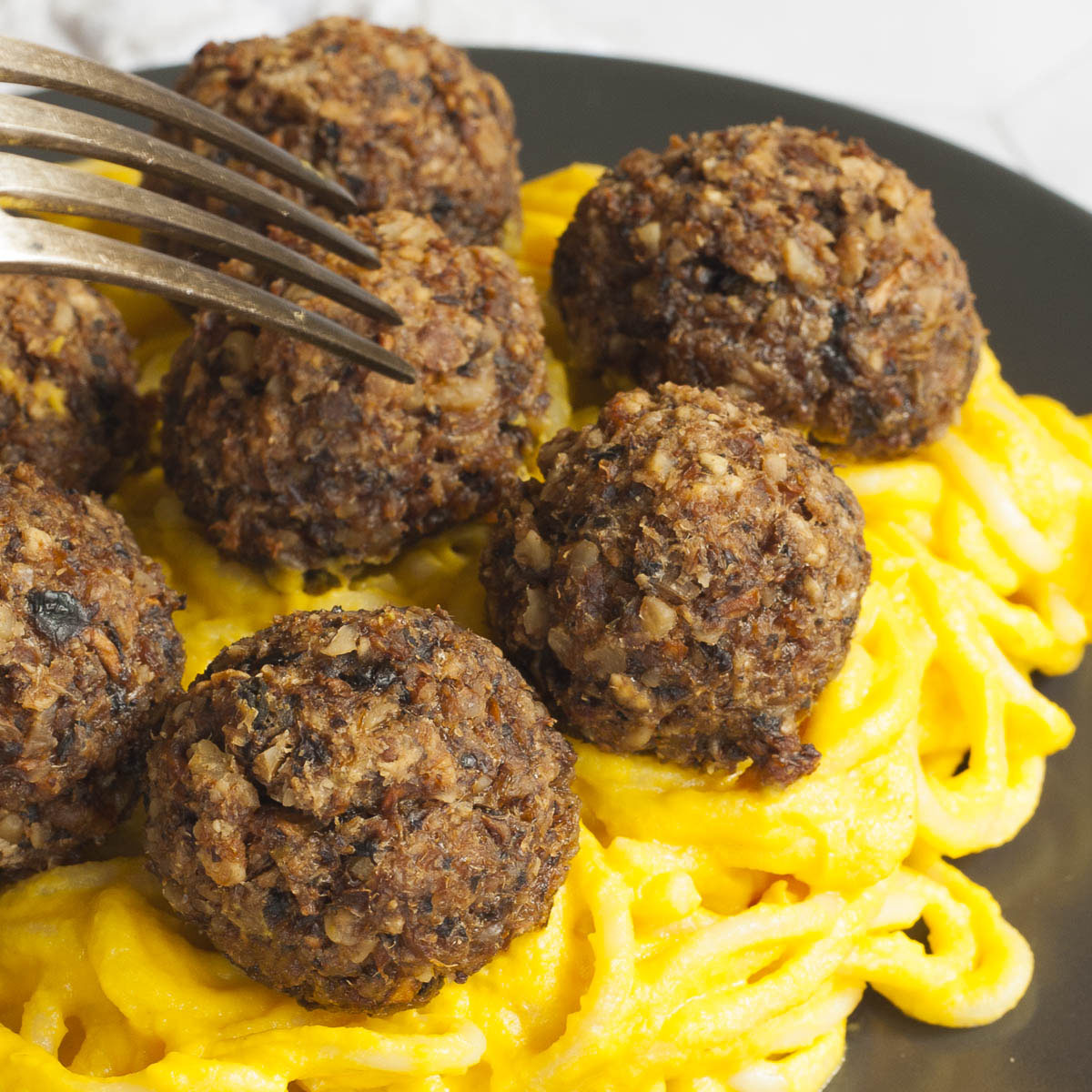 Several mushroom meatballs on top of spaghetti with yellow creamy sauce served on a black plate. A fork is taking on meatballs from the middle.