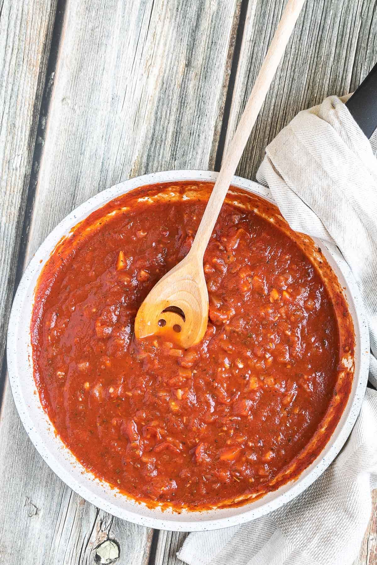 A white frying pan with red chunky sauce. A smiling wooden spatula is placed inside.