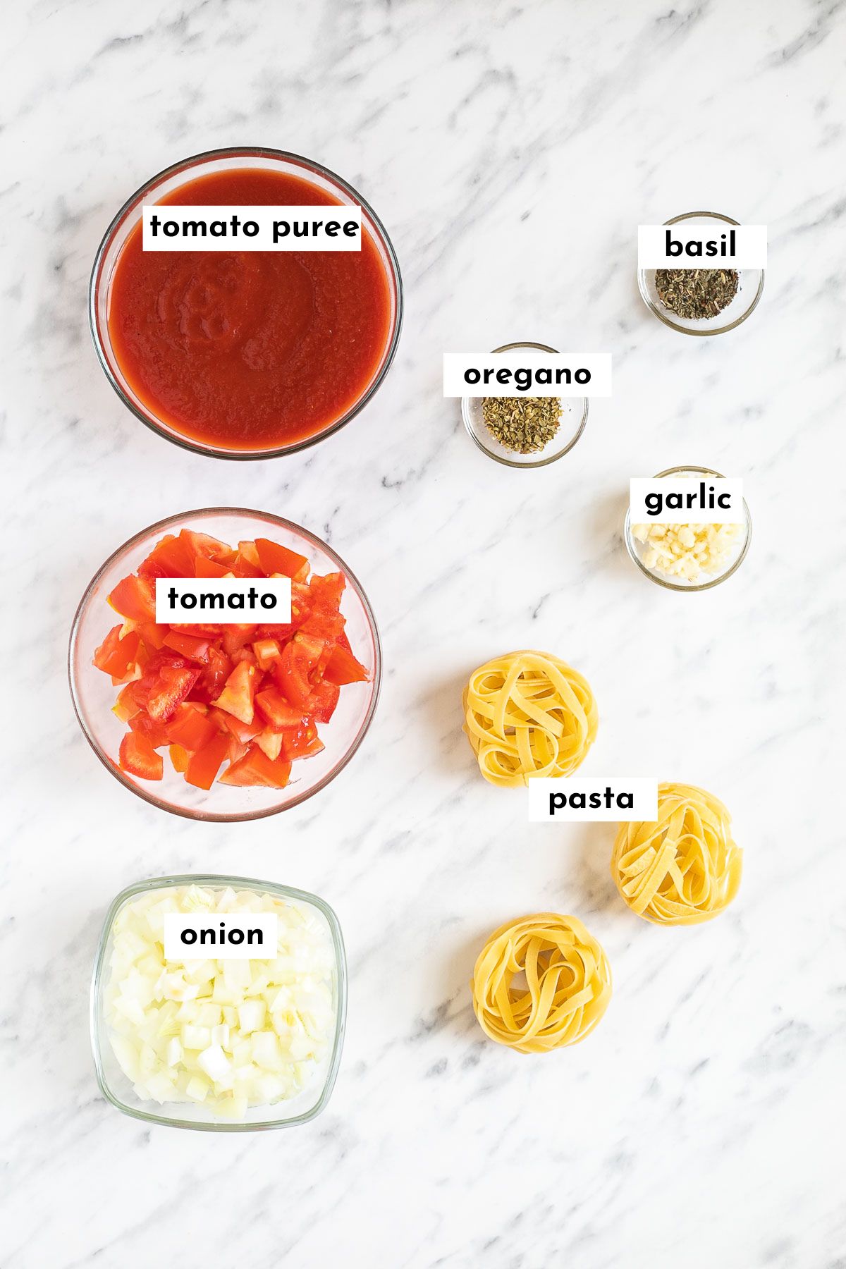 The ingredients of vegan marinara sauce is in small glass bowls tomato puree, chopped tomatoes, chopped onion, garlic, oregano, basil and dry pasta