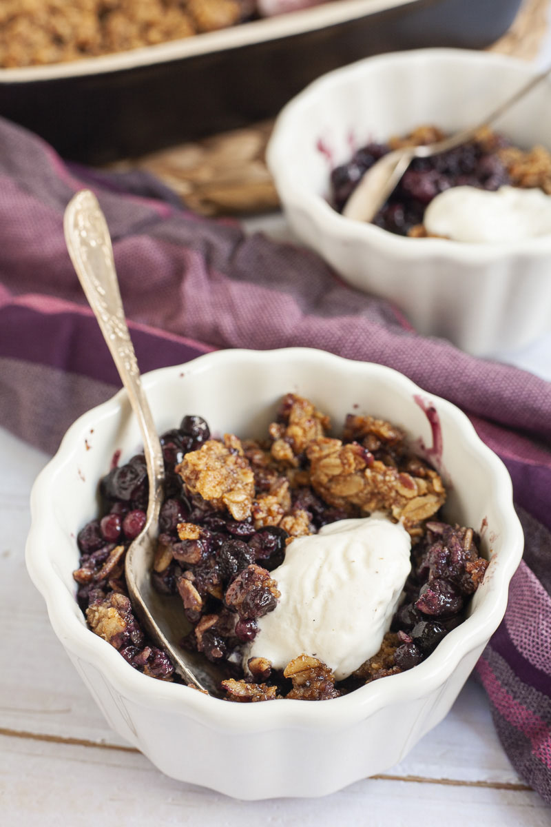 Up close a small white bowl with one serving of purple blueberries, golden brown crisps and a small scoop of white ice cream.