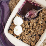 A spoon is in the middle of a white rectangle baking dish. Half of the dish is full of brown crisp, the other half is empty only the purple juices of the blueberry colors the bottom of the dish.