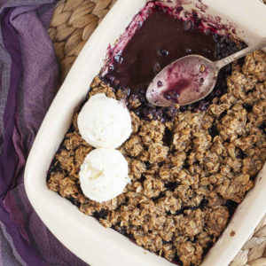 A spoon is in the middle of a white rectangle baking dish. Half of the dish is full of brown crisp, the other half is empty only the purple juices of the blueberry colors the bottom of the dish.