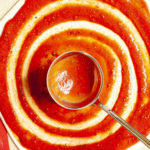 Red pizza sauce is spread in circles on top of a pizza crust with a ladle.