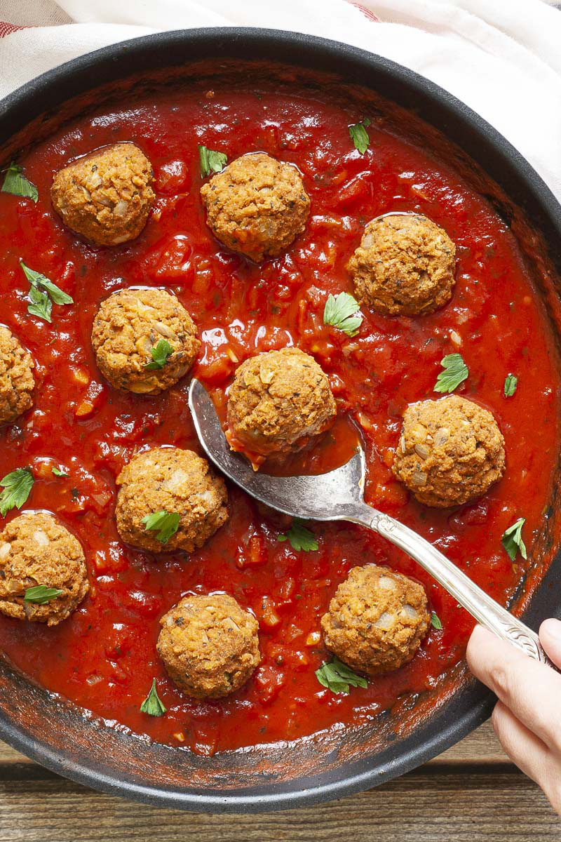 A skillet from above with several baked brown meatballs in bright red sauce and chopped green herbs. A spoon is taking one ball from the middle.