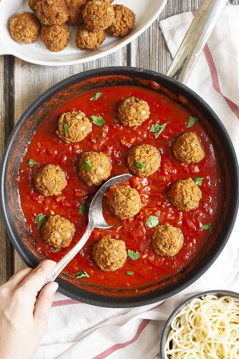 A skillet from above with several baked brown meatballs in bright red sauce and chopped green herbs. A spoon is taking one ball from the middle.