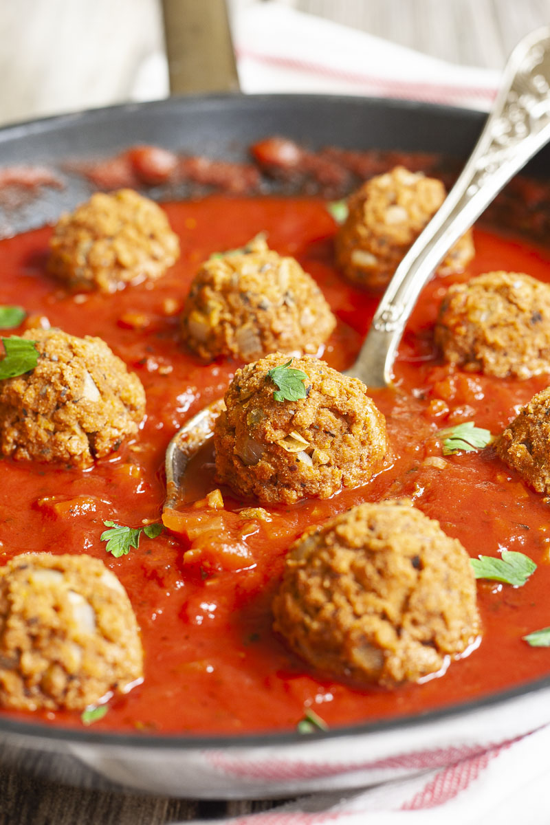 A skillet with several baked brown meatballs in bright red sauce and chopped green herbs. A spoon is taking one ball from the middle.