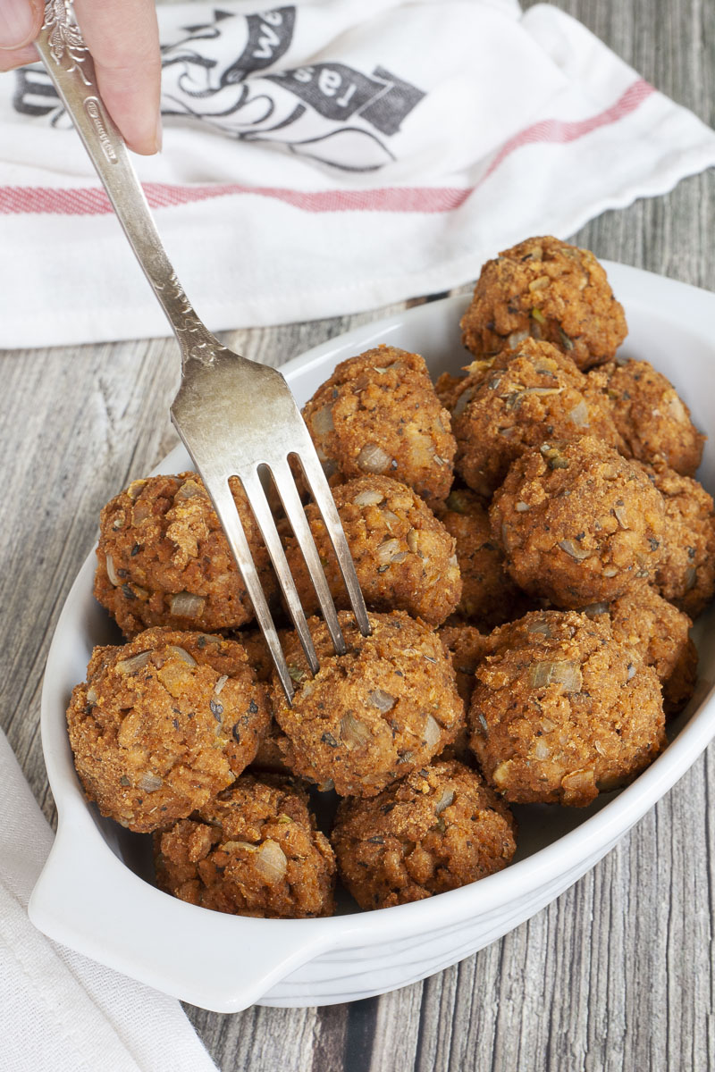 A white serving bowl full of baked brown vegan tvp meatballs. A fork is taking a ball from the middle.
