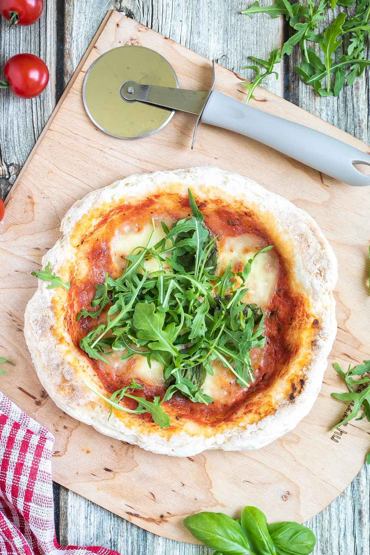 Pizza with fluffy crispy crust topped with tomato sauce, melted cheese and fresh arugula on a wooden board. Pizza cutter and more fresh herbs ingredients are around it. 