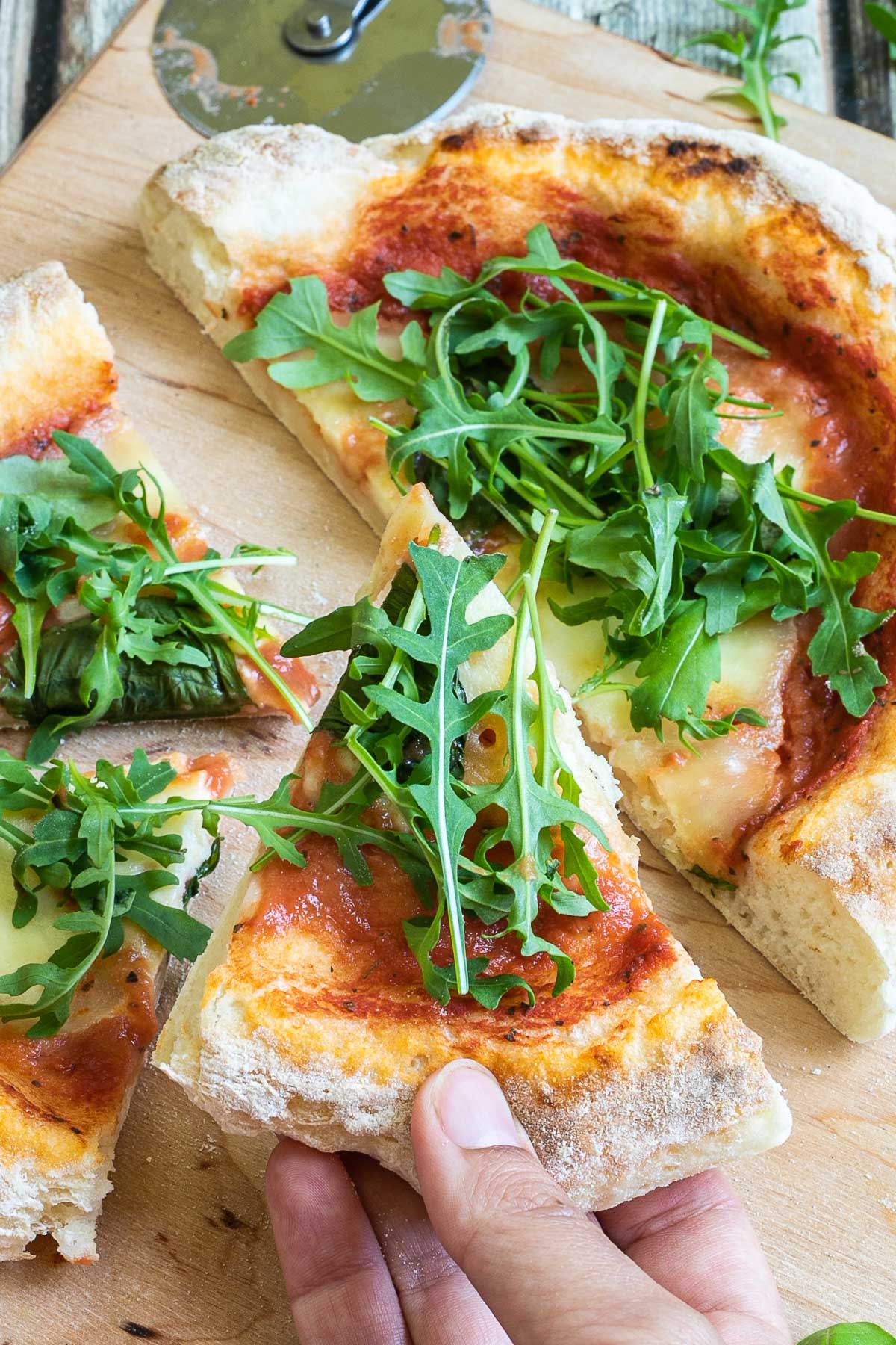 A pizza cut into slices with fluffy crispy crust topped with tomato sauce, melted cheese and fresh arugula on a wooden board. A hand is taking one slice.