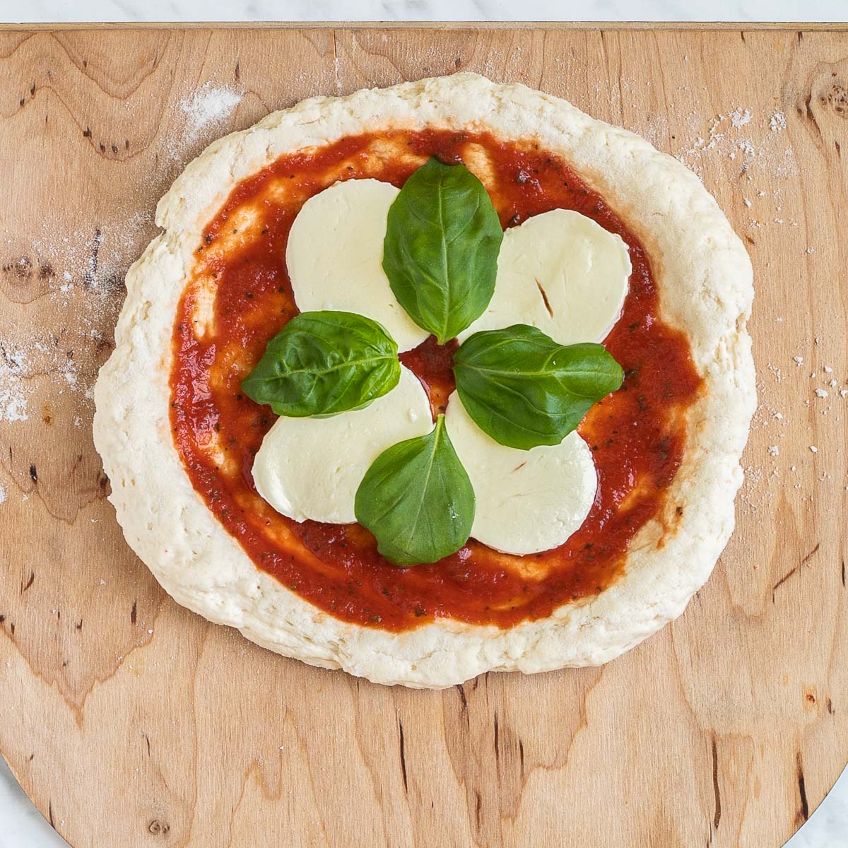 A pizza-shaped dough on a wooden pizza peel topped with tomato sauce, mozzarella and basil leaves.