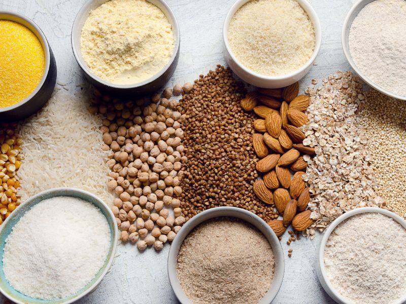 Many different flours in small bowls and the seeds, nuts and grains they are made of around them. 