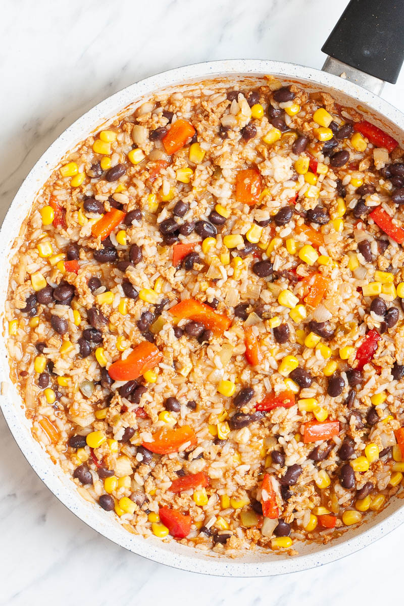 A white frying pan with colorful ingredients like black beans, corn, bell pepper strips and rice.