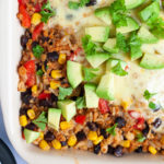 A ligth brown ceramic dish with colorful ingredients like black beans, corn, rice, melted cheese, bell pepper strips, chopped avocado, chopped green herbs.